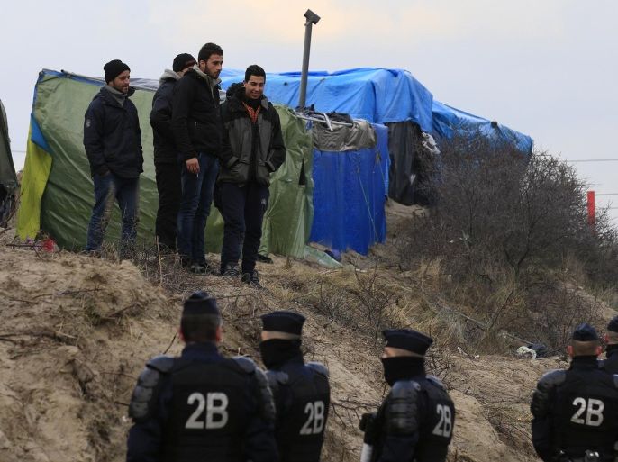 A group of migrants stand near tents as French riot policemen look on in the camp known as the "Jungle", a squalid sprawling camp in Calais, northern France, February 25, 2016. A French judge on Thursday upheld a government plan to partially demolish a shanty town for migrants trying to reach Britain on the outskirts of the northern port of Calais, an official spokesman said. REUTERS/Pascal Rossignol