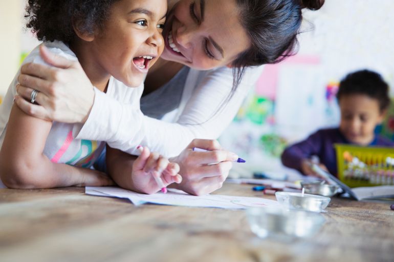 Affectionate, happy mother hugging toddler daughter coloring at table