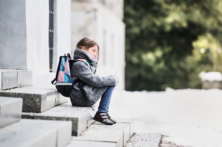 A portrait of a small girl with a schoolbag sitting on the stairs in front of school, waiting. كيف يؤثر عدم الذهاب إلى المدرسة على الأطفال؟