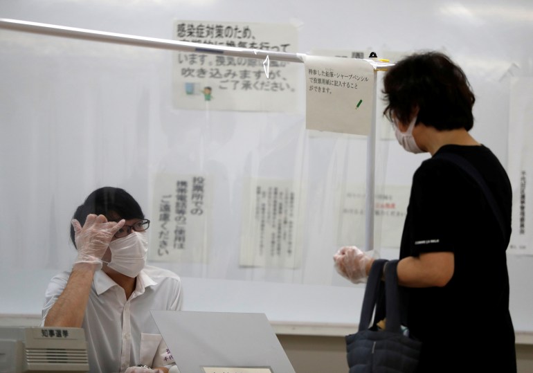 A member of Election Management Committee wearing a protective face mask and vinyl gloves talks with a voter behind a plastic curtain amid the coronavirus disease (COVID-19) outbreak, at a voting station for for the Tokyo Governor election in Tokyo