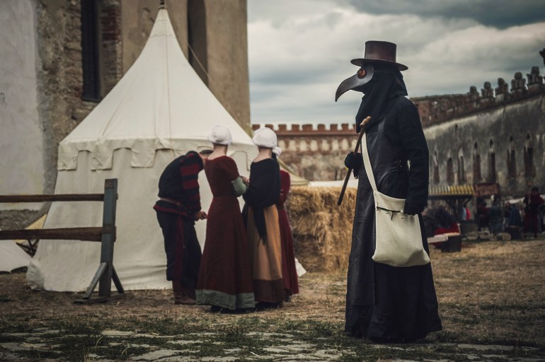 A masquerade historical scene reconstruction. Plague doctor in medieval old town. Castle and epidemic; Shutterstock ID 1683904612; purchase_order: ajnet; job: ; client: ; other: