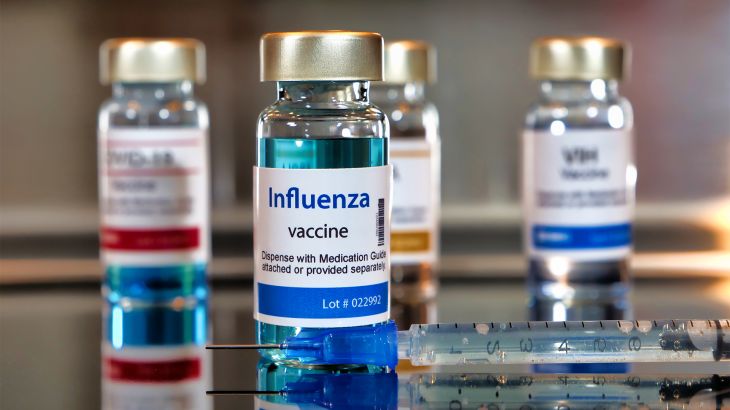 Bottles of influenza vaccine. ottles with a syringe on black table and stainless steel background. GettyImages-1250471564
