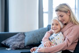A young Mother sits on a sofa at home, with her young daughter on her lap, between cancer treatments. She is hugging her tightly as they enjoy a close moment together. The little girl is dressed casually, has a head scarf on and is smiling.
