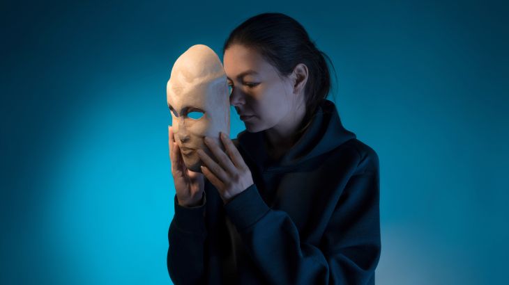 Hiding behind a mask, a young woman in a dark hoodie hides her face with a mask, self-identification problems and impostor syndrome. Portrait in the studio on a dark gray background.