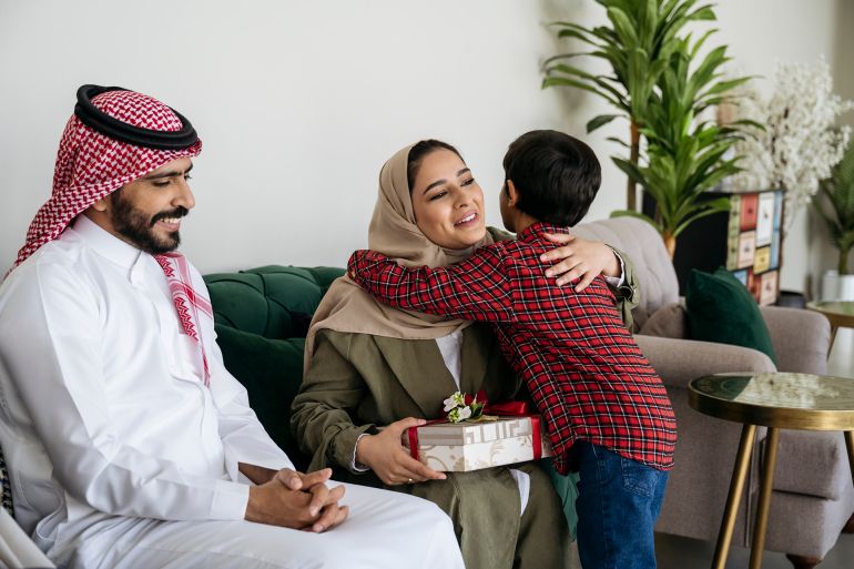 Smiling Saudi parents in traditional attire sitting on sofa in family home and enjoying hospitality and generosity expressed during holiday.